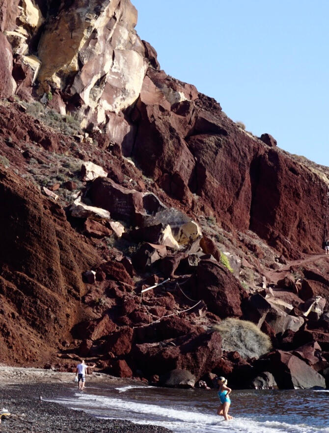 Wide-angle picture of large irregular maroon cliffs towering over a beach. Two people stand in the shallow water.