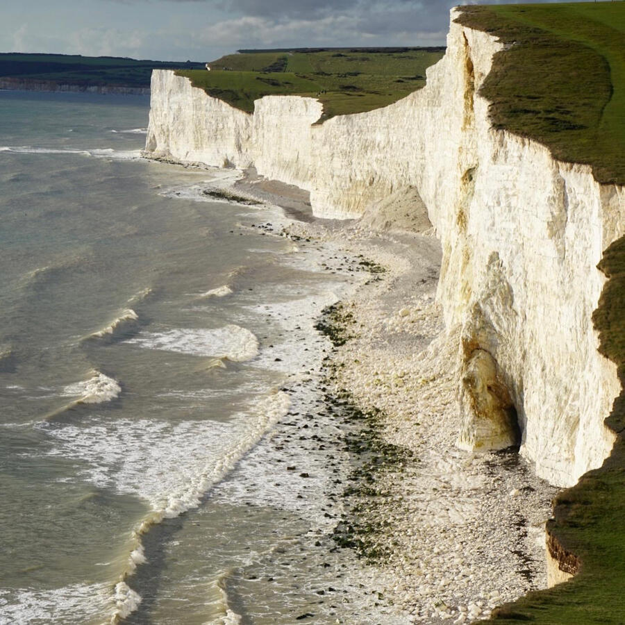 The view from the Seven Sisters hike on the coast of Eastbourne, East Sussex, ENG. White cliffs topped by greenery drop into a pale beach covered by gentle waves.